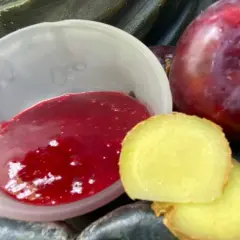 Eine Schale mit leuchtend roter asiatischer Pflaumensauce neben frischen Pflaumen und Ingwerscheiben.
