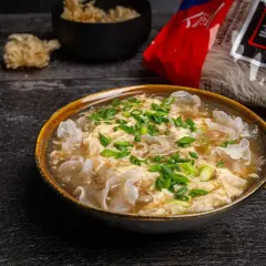 Eine dampfende Schüssel chinesische Suppe mit Ei, garniert mit Frühlingszwiebeln und Pilzen. Daneben liegt eine Packung Reisnudeln.