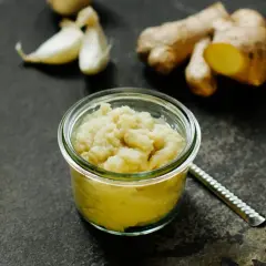 Frisch zubereitete ingwer knoblauch paste in einem Glas. Daneben liegen frische Knoblauchzehen und Ingwerwurzeln.