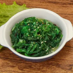 Schüssel mit leuchtend grünem Seetang Salat, garniert mit Sesam. Daneben ein Blatt grüner Salat.