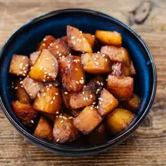 Glasierte Süßkartoffelwürfel mit Sesam, ein köstliches Beispiel für Banchan Rezepte, serviert in einer blauen Schüssel auf Holzuntergrund.