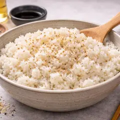 Eine Schüssel mit dampfendem, weißem Reis, bestreut mit Sesam. Daneben ein Holzstäbchen und kleine Schälchen mit Sojasauce und Öl. Ein Hauch von blauer Reis-Farbe könnte hier für eine Überraschung sorgen.