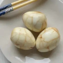 Drei marmorierte Tee Eier auf einer hellen Platte, daneben blaue Essstäbchen.