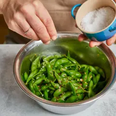 Hände würzen frisch gekochte Edamame mit Meersalz und Chiliflocken. So einfach ist es, Edamame zuzubereiten!