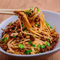 Ein Teller Dan Dan Noodles mit Hackfleisch, Erdnüssen und Frühlingszwiebeln, serviert mit Stäbchen.