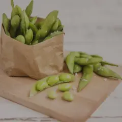 Frische Sojabohnen in einer Papiertüte und auf einem Holzbrett, bereit zum Zubereiten. Ein paar Bohnen sind aus der Hülse gefallen.