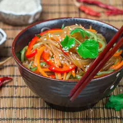Schüssel mit Japchae, einem koreanischen Nudelgericht, garniert mit Koriander und Sesam. Daneben Sojasauce, getrocknete Chilis und Reis.