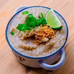 Herzhafter Reis Congee mit Rinderpansen, Frühlingszwiebeln, geröstetem Knoblauch und Limettenspalte in einer Schüssel auf Holzhintergrund.