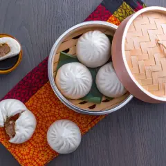 Dampfende chinesische Brote, gefüllt mit herzhaftem Fleisch, werden in einem Bambuskorb serviert. Eines ist aufgeschnitten und zeigt die köstliche Füllung.
