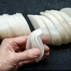 Eine Hand hält dünn geschnittene Rettichscheiben, bereit zum Einlegen. Weitere Scheiben liegen auf einem dunklen Untergrund.