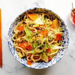 Schüssel mit leckeren Udon Nudeln kochen, garniert mit Gemüse, Frühlingszwiebeln und Sesam. Daneben liegen Essstäbchen und eine kleine Schale mit Chilis.