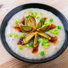 Ein cremiges Congee-Rezept mit geschnittenem konserviertem Ei und Frühlingszwiebeln, serviert in einer schwarzen Schüssel auf Holzhintergrund.