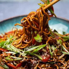 Ein Teller mit dampfendem Chow Mein, garniert mit Frühlingszwiebeln und Sesam. Essstäbchen heben eine Portion der Nudeln an.
