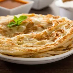 Ein Stapel knuspriger Roti Canai, garniert mit Minze, serviert auf einem Teller mit Curry und Joghurt im Hintergrund.