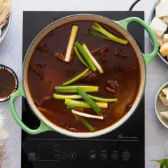 Ein köstlicher chinesischer Eintopf brodelt in einem grünen Topf, umgeben von Zutaten wie Pilzen, Tofu, Teigtaschen und Reisnudeln.