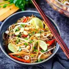 Schüssel mit asiatischem Glasnudelsalat, garniert mit Erdnüssen, Limettenspalten und Frühlingszwiebeln. Chopsticks liegen auf dem Teller.