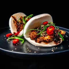 Zwei köstliche Bao Buns, gefüllt mit knusprigem Fleisch, garniert mit Chilis und Frühlingszwiebeln, auf einem schwarzen Teller.