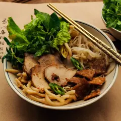 Cao Lầu: Nudelschüssel mit Schweinefleisch, Sprossen, Kräutern und knusprigen Krümeln. Goldene Stäbchen ruhen darauf.