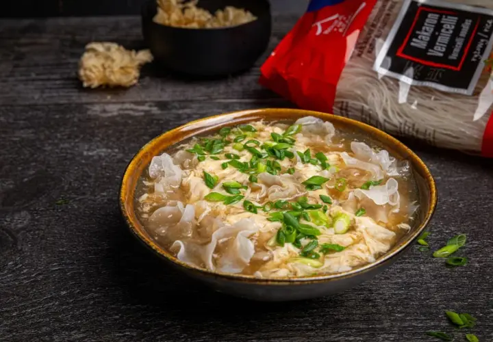 Eine dampfende Schüssel chinesische Suppe mit Ei, garniert mit Frühlingszwiebeln und Pilzen. Daneben liegt eine Packung Reisnudeln.
