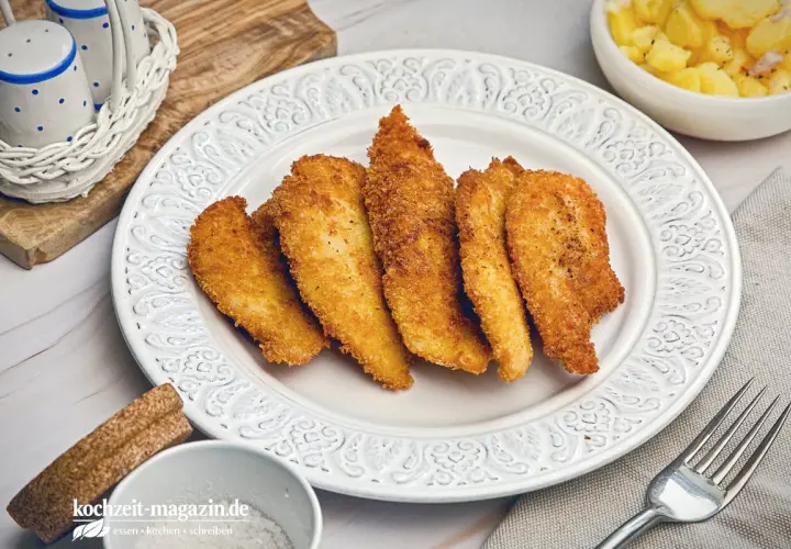 Knusprige Hähnchenstreifen in goldbrauner Panko Panade auf einem Teller, daneben Kartoffelsalat.