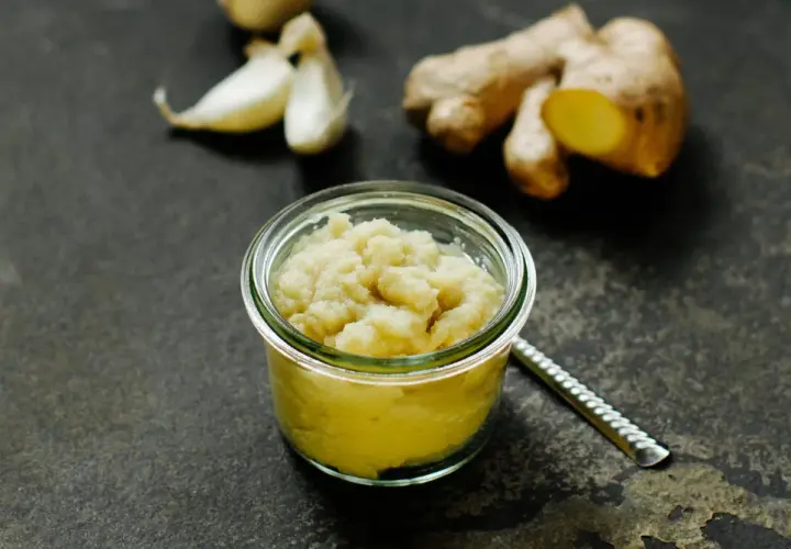 Frisch zubereitete ingwer knoblauch paste in einem Glas. Daneben liegen frische Knoblauchzehen und Ingwerwurzeln.
