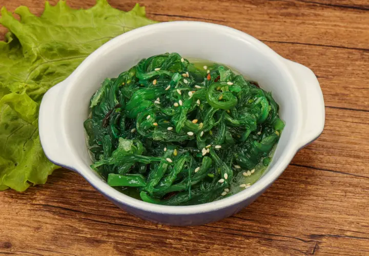 Schüssel mit leuchtend grünem Seetang Salat, garniert mit Sesam. Daneben ein Blatt grüner Salat.