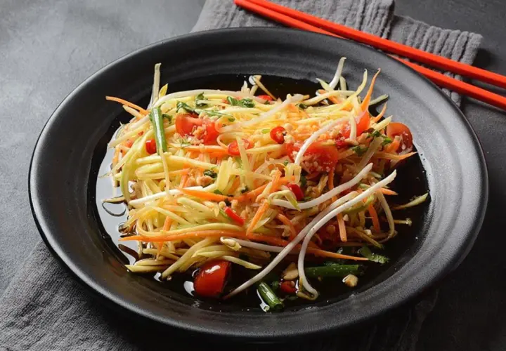 Ein bunter Papaya Salat mit Karottenstreifen, Kirschtomaten und Sprossen, serviert auf einem schwarzen Teller mit roten Stäbchen.