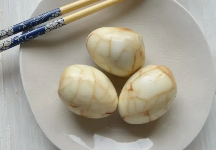 Drei marmorierte Tee Eier auf einer hellen Platte, daneben blaue Essstäbchen.