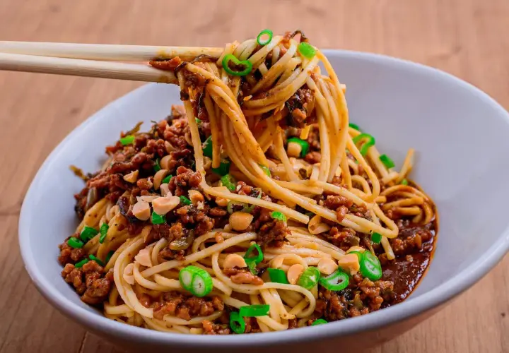 Ein Teller Dan Dan Noodles mit Hackfleisch, Erdnüssen und Frühlingszwiebeln, serviert mit Stäbchen.