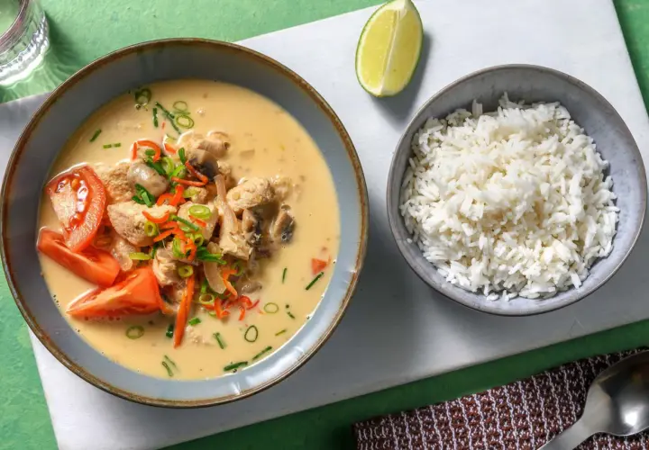 Khao tom: Eine Schüssel mit cremigem Hühner-Kokos-Eintopf, garniert mit Tomaten und Frühlingszwiebeln, daneben eine Schüssel Reis und eine Limettenspalte.