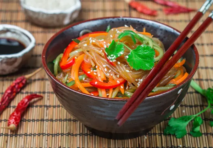 Schüssel mit Japchae, einem koreanischen Nudelgericht, garniert mit Koriander und Sesam. Daneben Sojasauce, getrocknete Chilis und Reis.