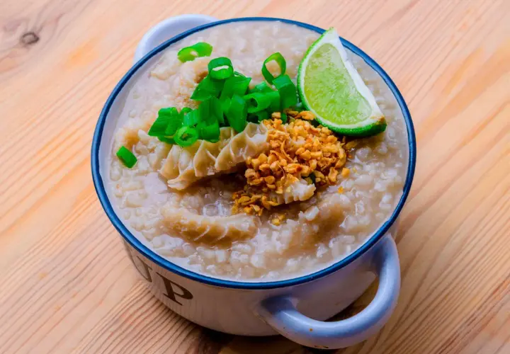 Herzhafter Reis Congee mit Rinderpansen, Frühlingszwiebeln, geröstetem Knoblauch und Limettenspalte in einer Schüssel auf Holzhintergrund.