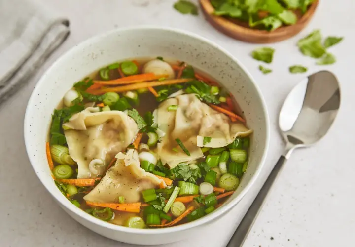 Eine Schüssel mit dampfender Suan La Tang, gefüllt mit Teigtaschen, Karottenstreifen und Frühlingszwiebeln.