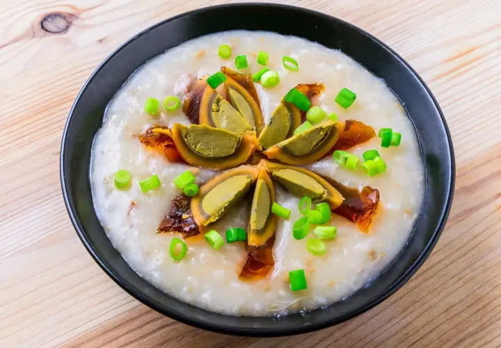 Ein cremiges Congee-Rezept mit geschnittenem konserviertem Ei und Frühlingszwiebeln, serviert in einer schwarzen Schüssel auf Holzhintergrund.