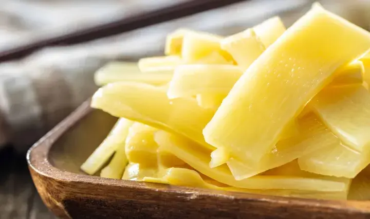 Holzschale mit leuchtend gelben, geschnittenen Bambussprossen, die gesund und appetitlich aussehen. Ein dunkler Löffel liegt im Hintergrund.