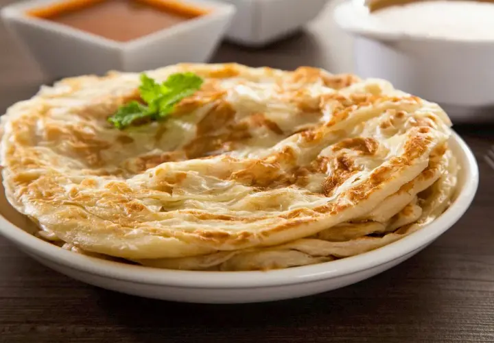 Ein Stapel knuspriger Roti Canai, garniert mit Minze, serviert auf einem Teller mit Curry und Joghurt im Hintergrund.