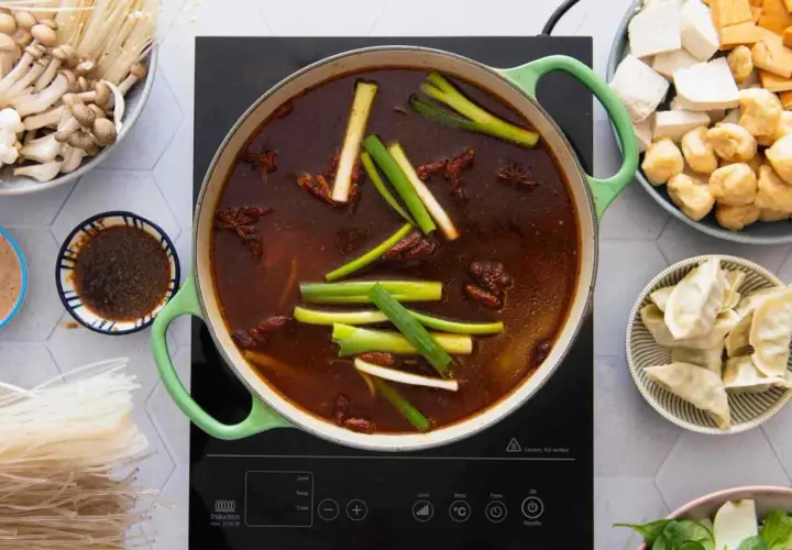 Ein köstlicher chinesischer Eintopf brodelt in einem grünen Topf, umgeben von Zutaten wie Pilzen, Tofu, Teigtaschen und Reisnudeln.
