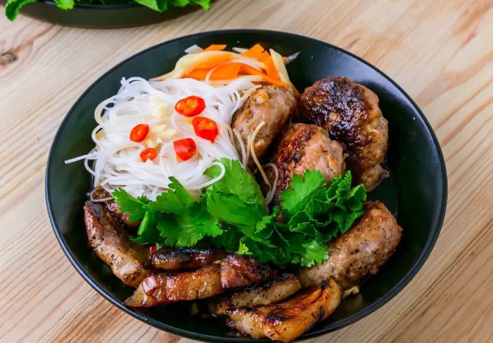 Bun cha Hanoi: gegrilltes Schweinefleisch und Fleischbällchen mit Reisnudeln, Kräutern und Chili auf einem Teller.