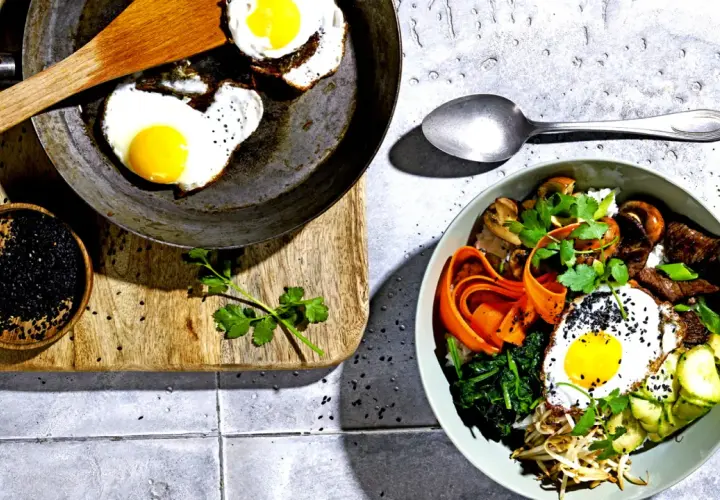 Ein köstliches Bibimbap-Rezept: Bunte Schüssel mit Reis, Gemüse, Rindfleisch, Pilzen und Spiegelei, garniert mit schwarzen Sesamkörnern.