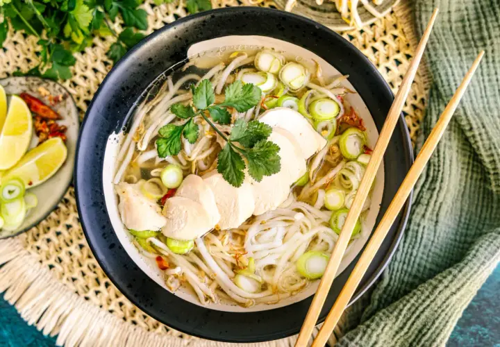 Eine dampfende Schüssel Pho Ga mit zarten Hühnerbrustscheiben, Reisnudeln, Sojasprossen und Frühlingszwiebeln. Dazu Limettenspalten und Chili.