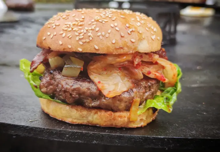 Ein saftiger korean burger mit Kimchi, Gurken und Speck auf einem Sesambrötchen.