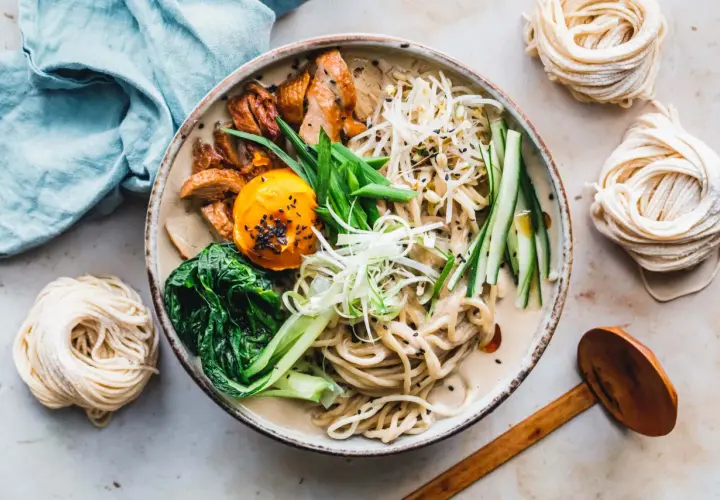 Schüssel mit cremiger Ramen-Suppe, Hühnchen, Ei, Gemüse und frischen **Rahmen Nudeln** daneben.