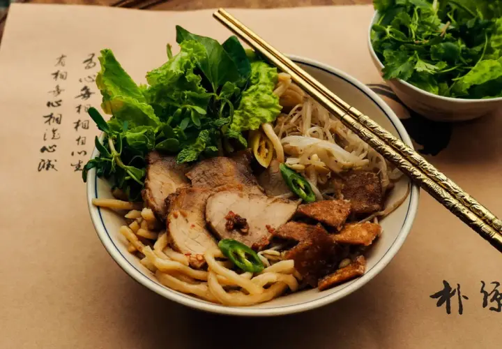 Cao Lầu: Nudelschüssel mit Schweinefleisch, Sprossen, Kräutern und knusprigen Krümeln. Goldene Stäbchen ruhen darauf.