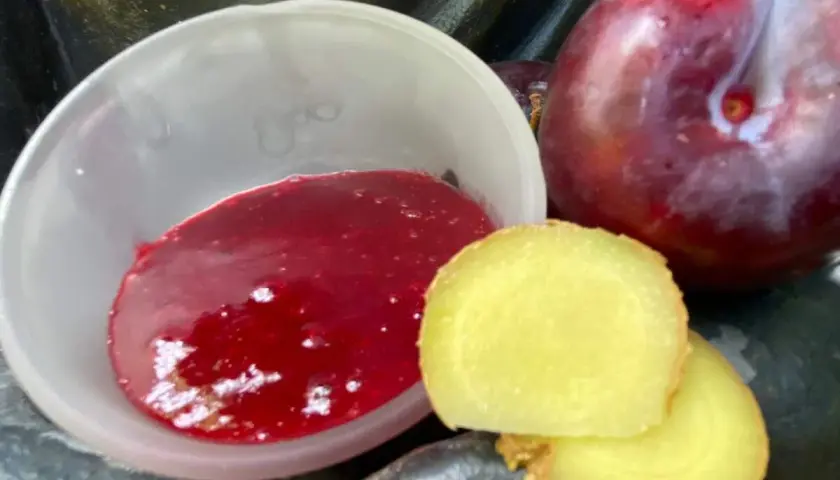 Eine Schale mit leuchtend roter asiatischer Pflaumensauce neben frischen Pflaumen und Ingwerscheiben.