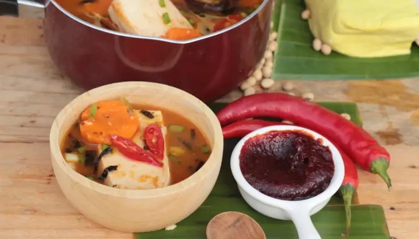 Gochujang Zutaten: Ein Topf mit Tofu-Eintopf, eine Schüssel mit scharfem Eintopf, rote Chilis, Sojabohnen und ein Block Tofu auf Bananenblättern.