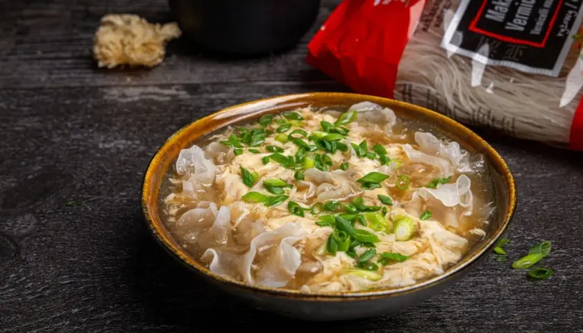 Eine dampfende Schüssel chinesische Suppe mit Ei, garniert mit Frühlingszwiebeln und Pilzen. Daneben liegt eine Packung Reisnudeln.