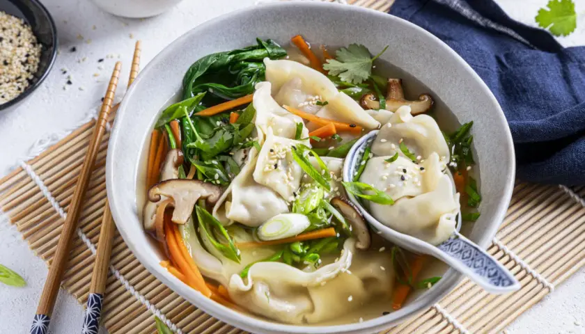Eine dampfende Schüssel **Wantan Suppe** mit zarten Teigtaschen, Shiitake-Pilzen, Karotten und Frühlingszwiebeln, garniert mit Sesam.