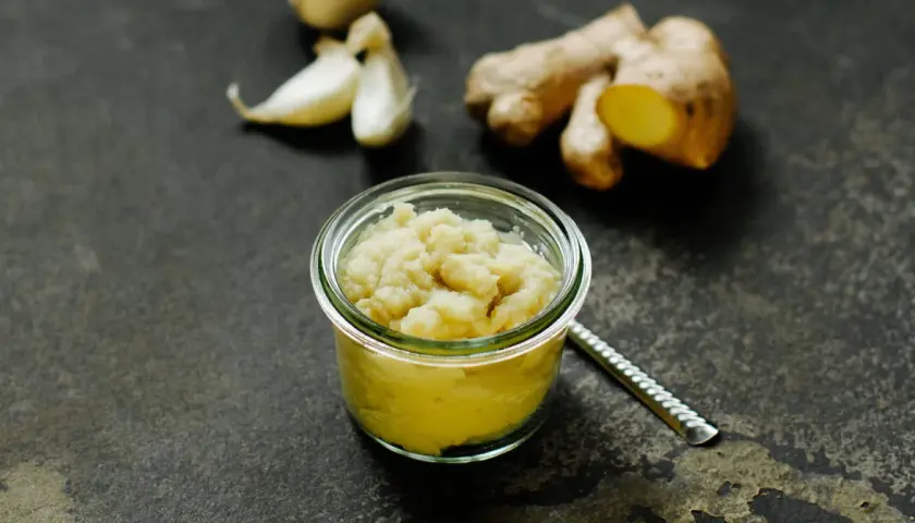 Frisch zubereitete ingwer knoblauch paste in einem Glas. Daneben liegen frische Knoblauchzehen und Ingwerwurzeln.