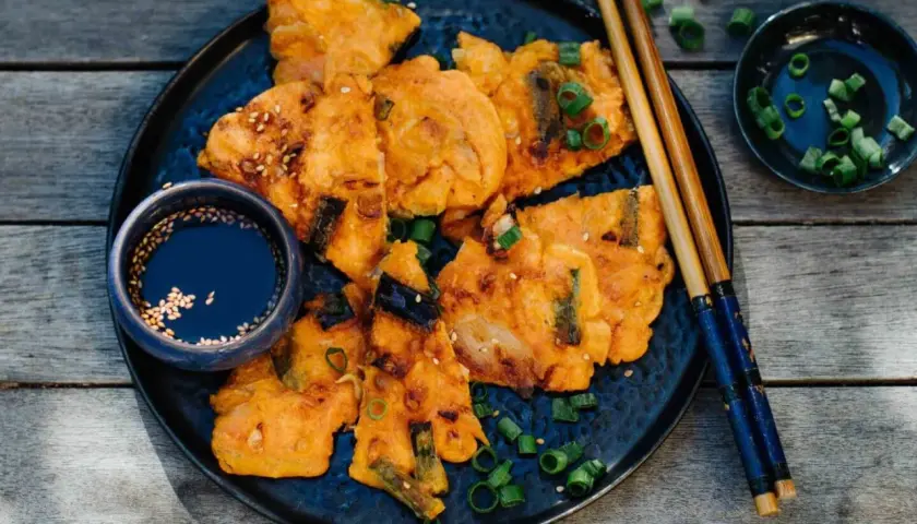 Knusprige Kimchi-Pfannkuchen mit Frühlingszwiebeln und Sesam, serviert mit Dip und Stäbchen.