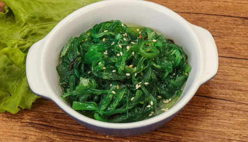 Schüssel mit leuchtend grünem Seetang Salat, garniert mit Sesam. Daneben ein Blatt grüner Salat.